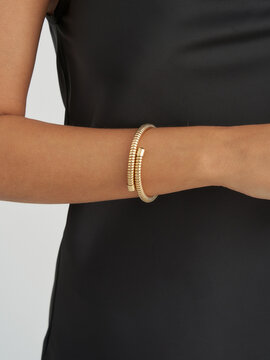 A person's arm wearing a stylish gold bracelet with a textured pattern, complemented by a sleek black dress. The bracelet is prominently featured, highlighting its elegance