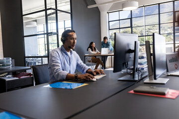 In office, Asian man using computer with headset, colleagues collaborating nearby