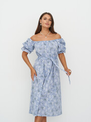 Woman stands in blue, floral, off-the-shoulder dress with puffy sleeves and tied waist. She has...