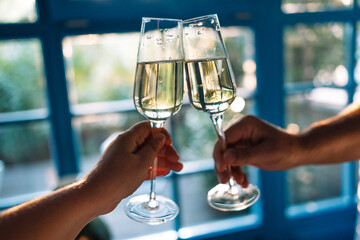Male and female hands hold out glasses of delicious champagne for a toast at home. A woman and a man clink goblets with sparkling wine against the background of windows with blue frames.