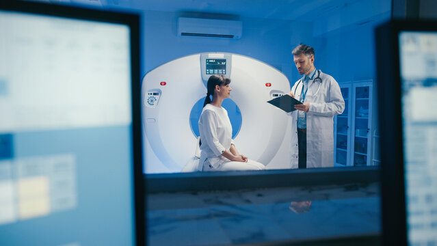 Pale woman sitting still on scanner bed and looking at doctor with composed expression. Medical specialist reviewing patient data and getting ready to starting scanning process in clinical room.