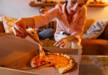 A pizza and a girl taking a piece from a cardboard box. A student in pajamas eats fast food at home in the evening.
