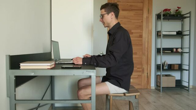 Young man working remotely in business shirt and underwear during video call at home showing humorous side of remote freelancing. Self-employed man, online work.