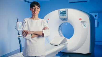 Pretty Caucasian woman crossing arms and smiling near CT machine in clean medical room. Standing...