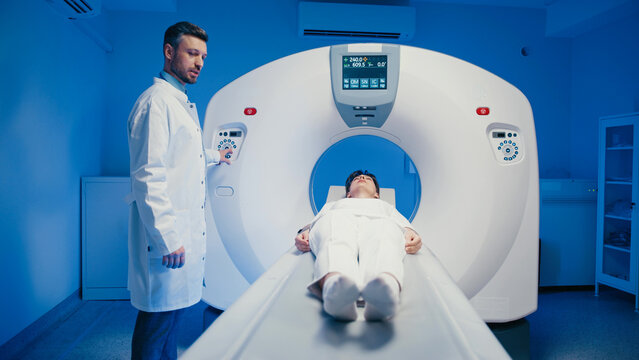 Caucasian male doctor wearing white lab coat while standing near MRI scanner and adjusting settings. Focused technician preparing medical imaging while patient lying inside large diagnostic machine.