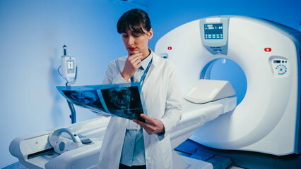 Focused female medical specialist examining brain scans near diagnostic imaging machine. Wearing...