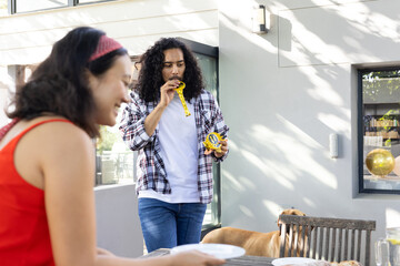 Celebrating outdoors, man blowing party horn while woman sets table with plates