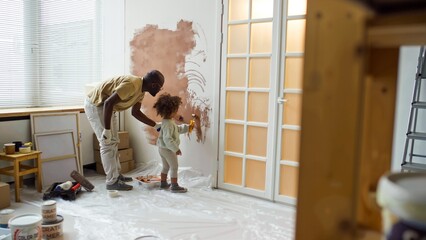 African American father painting wall with young daughter in light-filled room with scattered...