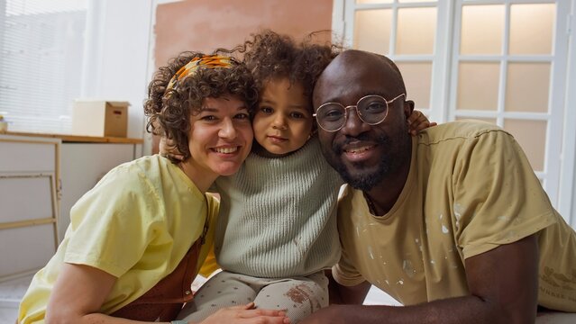 Portrait of biracial family sitting on couch, smiling warmly at camera. Family includes curly-haired woman, child with curly hair, and man with glasses wearing khaki shirt