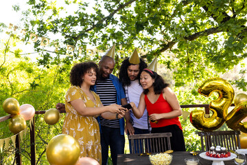 Fototapeta premium Friends celebrating birthday outdoors, wearing party hats and looking at smartphone