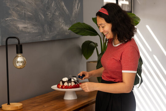Naklejki Asian woman decorating cake with strawberries at home, smiling and enjoying process