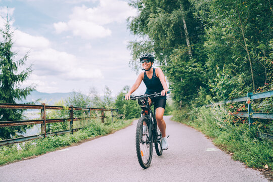 Portrait of happy smiling woman dressed in cycling clothes, helmet and sunglasses riding bicycle on asphalt out-of-town bicycle path. Active sporty people concept image