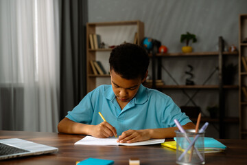 Naklejka premium Young african american child boy studying at home, kid sits at desk attends school class online on laptop, listening lesson and writing information in notebook.