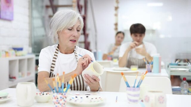 Interested elderly woman attending pottery class, enthusiastically painting on ceramic cup, creating unique custom design. Creative hobby concept