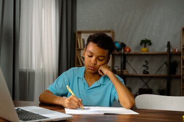 Young african american child boy studying at home, kid sits at desk attends school class online on laptop, listening lesson and writing information in notebook.