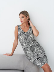 Woman in sleeveless black and white zebra-patterned dress sits on gray sofa, touching hair with one...