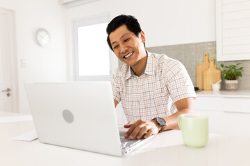 Smiling man using laptop at home, enjoying coffee in bright kitchen