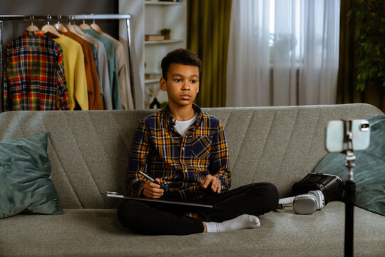 Young african american child boy spending time at home, kid blogger sits on sofa records video on smartphone, talking at camera using graphic tablet and gives review.