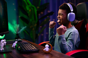 Young african american child boy spending time at home, kid streamer gamer in headphones sits at...