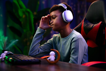 Young african american child boy spending time at home, kid streamer gamer in headphones sits at...