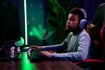 Young african american child boy spending time at home, kid streamer gamer in headphones sits at...
