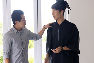 Proud father congratulating son in graduation gown at home, sharing joyful moment