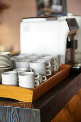 coffee cups stacked near espresso machine in cozy café setting. wood tray elegantly holds sleek white cups, creating inviting atmosphere perfect for coffee enthusiasts and baristas.