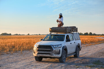 Woman on rooftop tent vehicle in a dry savannah, golden light, ready for wildlife photography. Clean composition with wide, open savannah and pastel sky.
