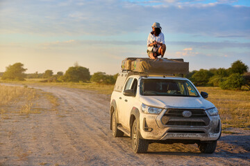 Woman on rooftop tent vehicle in a dry savannah, golden light, ready for wildlife photography. Clean composition with wide, open savannah and pastel sky.