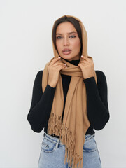 A woman with long dark hair wears a light brown headscarf and black sweater, holding the scarf with...