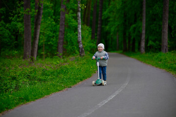 Obraz premium Little boy riding a green scooter on a paved path through a lush forest. Child dressed in a vest, beanie, and sneakers enjoying outdoor playtime in nature. Fun and active childhood moment.