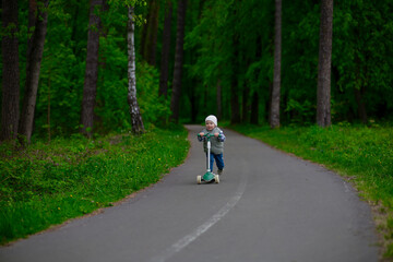 Fototapeta premium Little boy riding a green scooter on a paved path through a lush forest. Child dressed in a vest, beanie, and sneakers enjoying outdoor playtime in nature. Fun and active childhood moment.