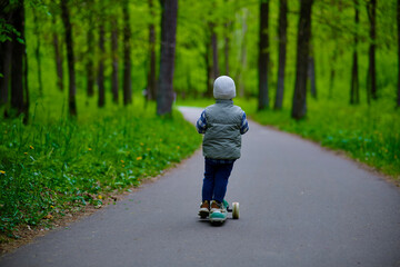 Fototapeta premium Little boy riding a green scooter on a paved path through a lush forest. Child dressed in a vest, beanie, and sneakers enjoying outdoor playtime in nature. Fun and active childhood moment.