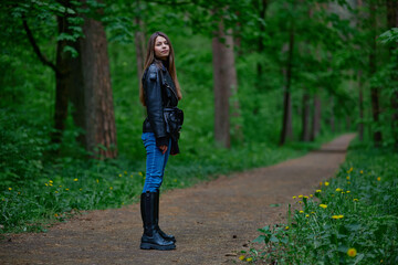 Obraz premium Confident young woman standing on a forest path, wearing a stylish black leather jacket, jeans, and black boots, surrounded by lush green trees in a serene natural setting.
