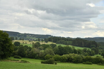 Fototapeta premium Paysage rural et champêtre entre collines boisées et prairies sous l'ombre des nuages à Bra (Lierneux)
