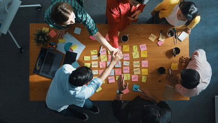Top view of diverse business team putting hand together and clapping hand while brainstorming marketing idea or creative startup project by using sticky notes and stick at meeting table. Convocation.