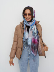 Woman wearing brown puffer jacket, colorful scarf, sunglasses, and blue jeans poses while adjusting...
