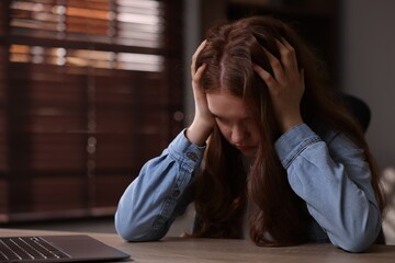Sad teenage girl with laptop at table indoors