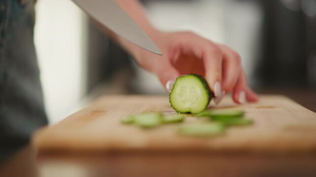 Close up of person slicing cucumber into four neat sections on wooden cutting board, with soft blur background and detailed view of hand, knife blade, and freshly cut green vegetable