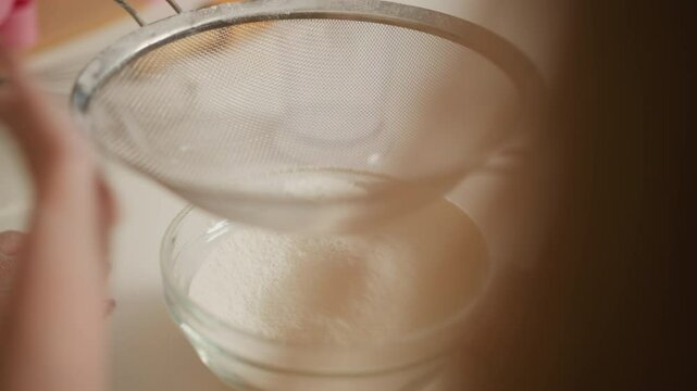 close up of flour gently being sifted through fine mesh sieve into glass bowl on white kitchen surface, hands nearby with soft light highlighting smooth texture of flour in calm baking