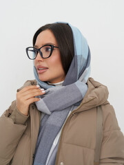 Woman with glasses wears a light blue and grey scarf and a beige jacket. Her dark hair is partially...