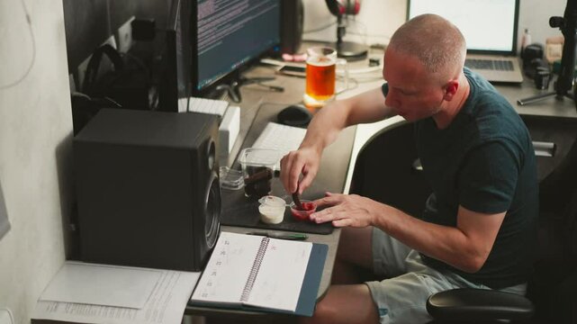 portrait view of remote worker dipping dark bread snack into sauce at home office desk with cup of beer beside computer monitor, keyboard, mouse, and opened notebook planner on wooden table