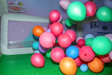Colorful balloons of various sizes are scattered across a green play area, enclosed by a netted fence. Bright hues of pink, blue, green, and orange are prominently featured