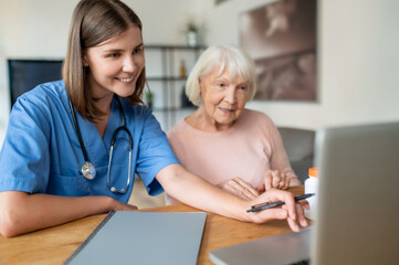 Female nurse in uniform using laptop with elderly patient in offering support for senior woman