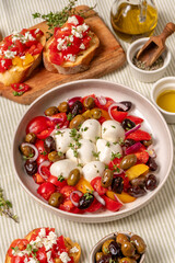 Fresh Italian style antipasto salad with colorful tomatoes, mozzarella balls, and a variety of olives, garnished with herbs, served in a bowl on a textured tablecloth with olive oil and bread 