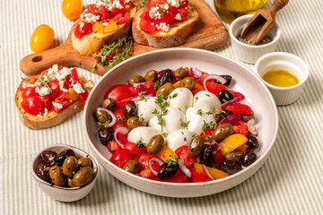 Fresh Italian style antipasto salad with colorful tomatoes, mozzarella balls, and a variety of olives, garnished with herbs, served in a bowl on a textured tablecloth with olive oil and bread 