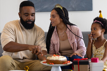 Father cutting birthday cake at home, daughter watching excitedly with family