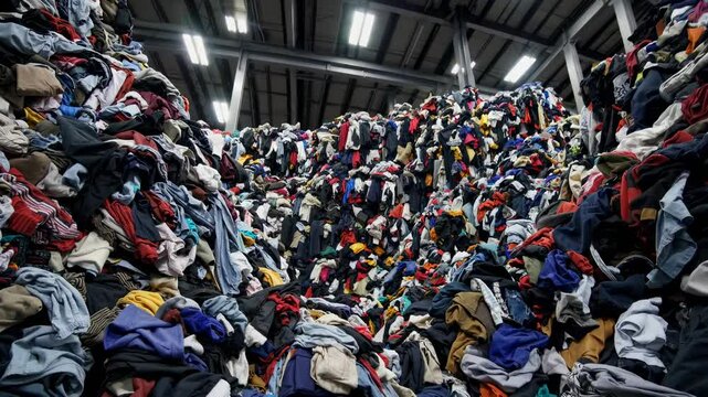 Low-angle shot of massive piles of colorful clothes in a warehouse, highlighting the scale of textile waste. Industrial video style.