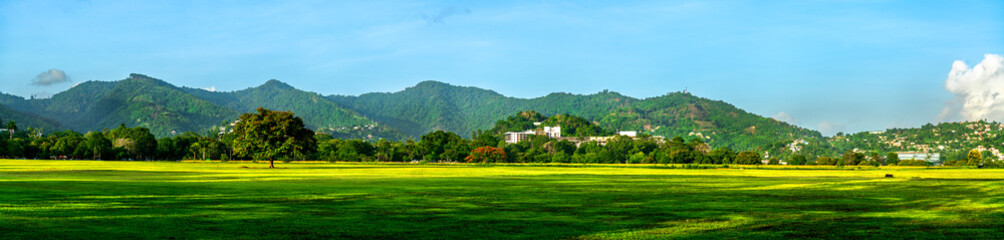 A wide panoramic view of Queen's Park Savannah in Port of Spain, Trinidad, featuring a vast green field bordered by trees and backed by the lush Northern Range mountains under a clear sky.
