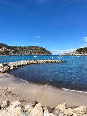 The arched pier goes out into the bay of the gulf. Behind it are yachts at anchor in the sea bay and yellow buoys. On a high cliff is a white lighthouse. The island of Mallorca. Spain